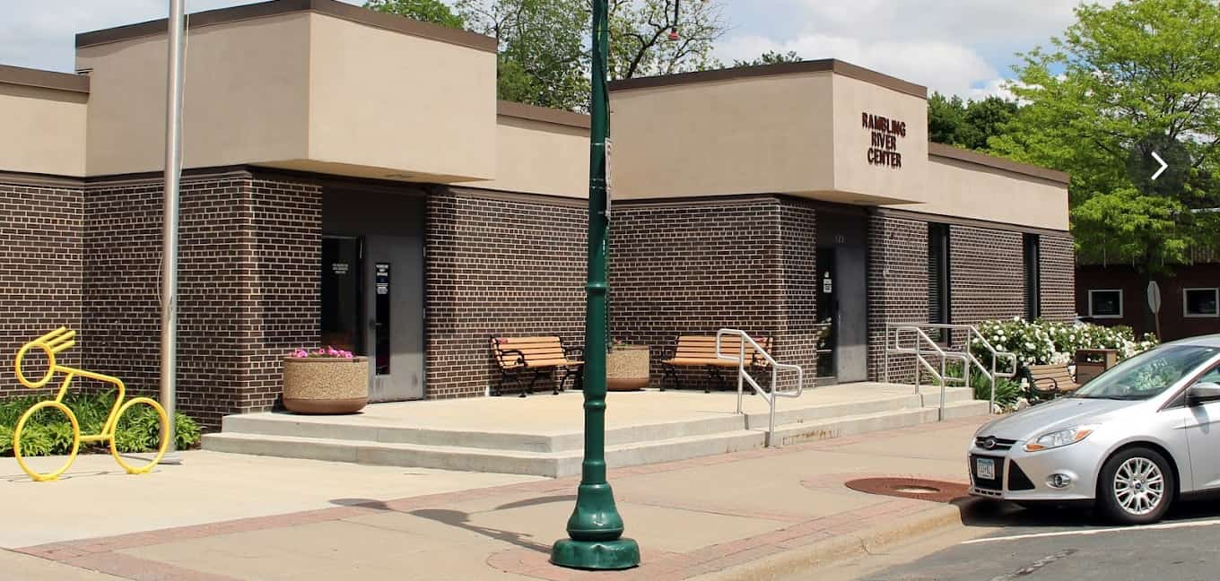 Exterior view of the Rambling River Center with benches, a bike rack sculpture, and a green lamppost in front of the entrance on a sunny day